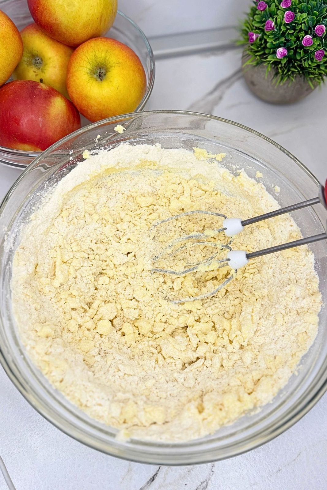 Crumbly dough for apple pie bars in a glass bowl with electric mixer beaters.