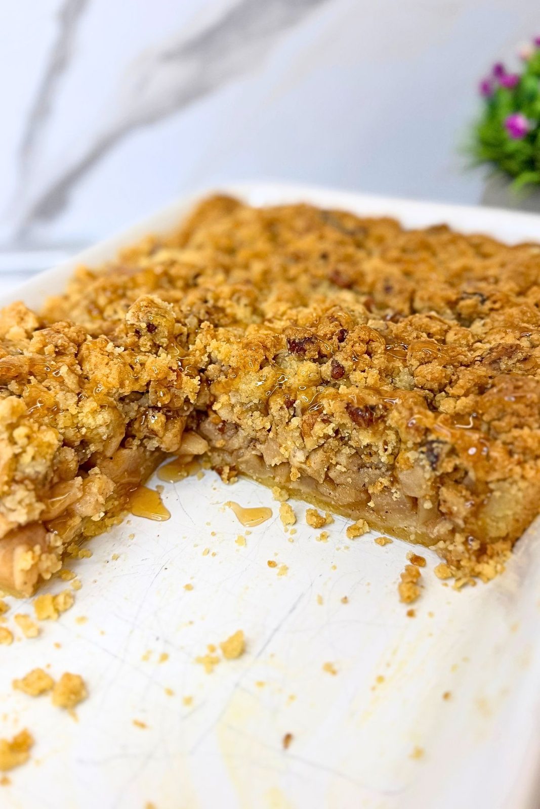 Apple pie bars with walnut crumble and caramel drizzle in a white baking dish, with one piece removed to reveal the apple filling and buttery crust.