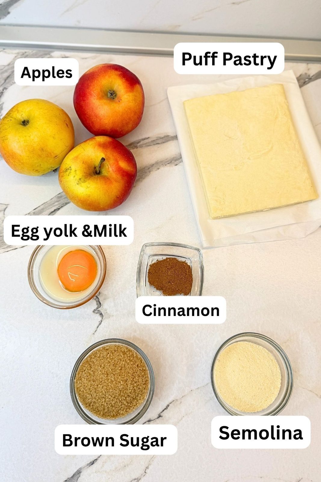 Ingredients for apple turnovers: puff pastry, apples, brown sugar, cinnamon, semolina, egg yolk, and milk arranged on a marble countertop.