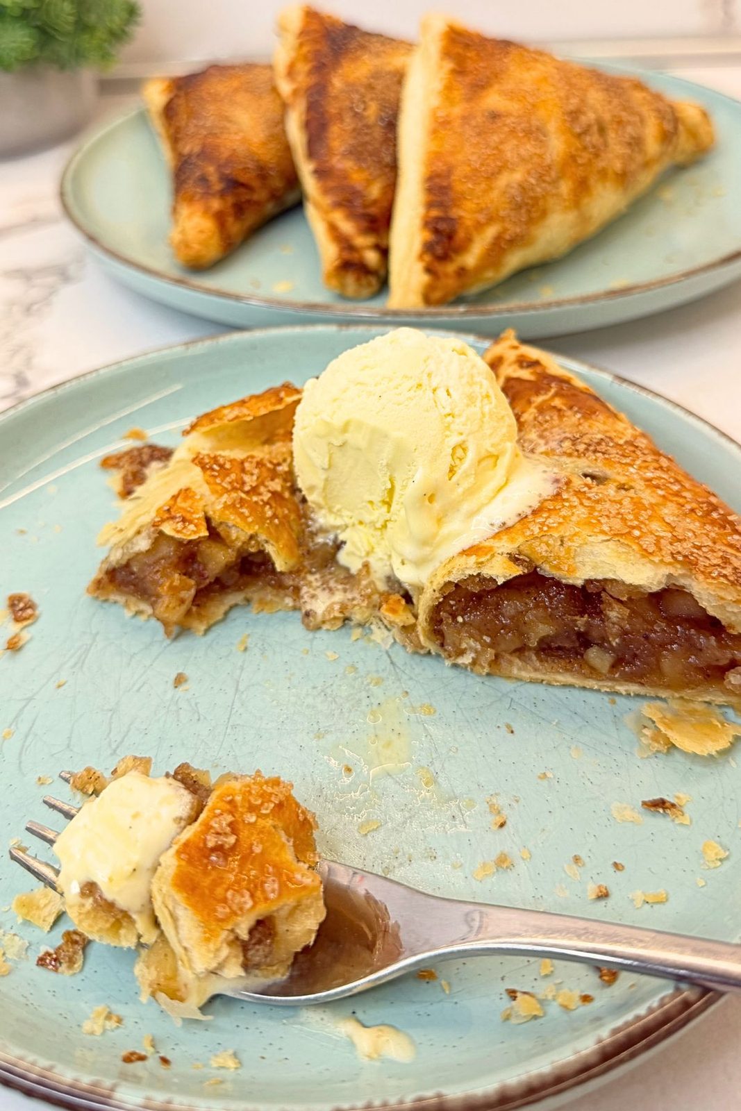 Apple turnover with cinnamon apple filling and vanilla ice cream on a blue plate, with a bite of flaky pastry and ice cream on a fork.