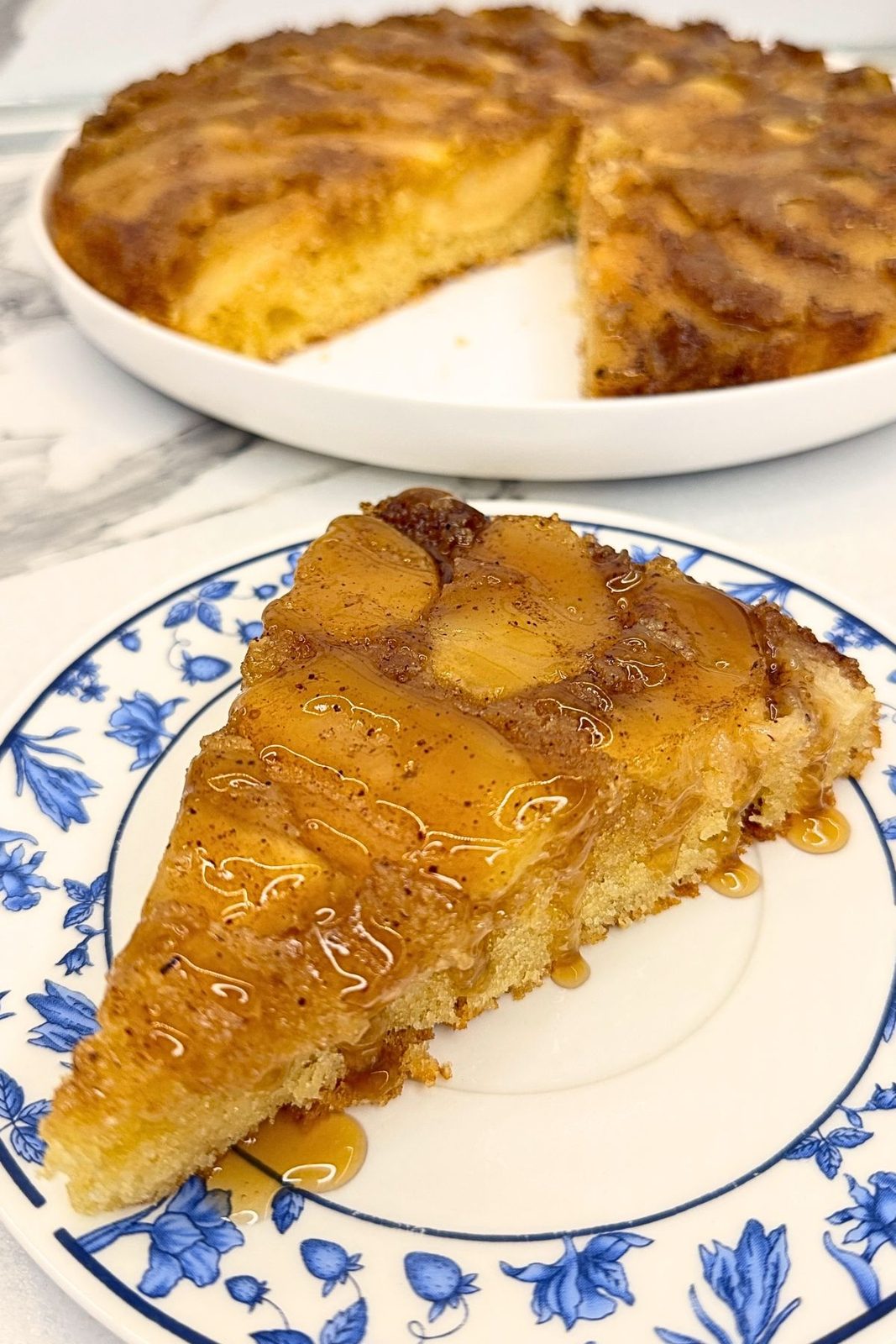 Slice of apple upside down cake with caramelized apples on a blue-and-white plate, with the remaining cake in the background.