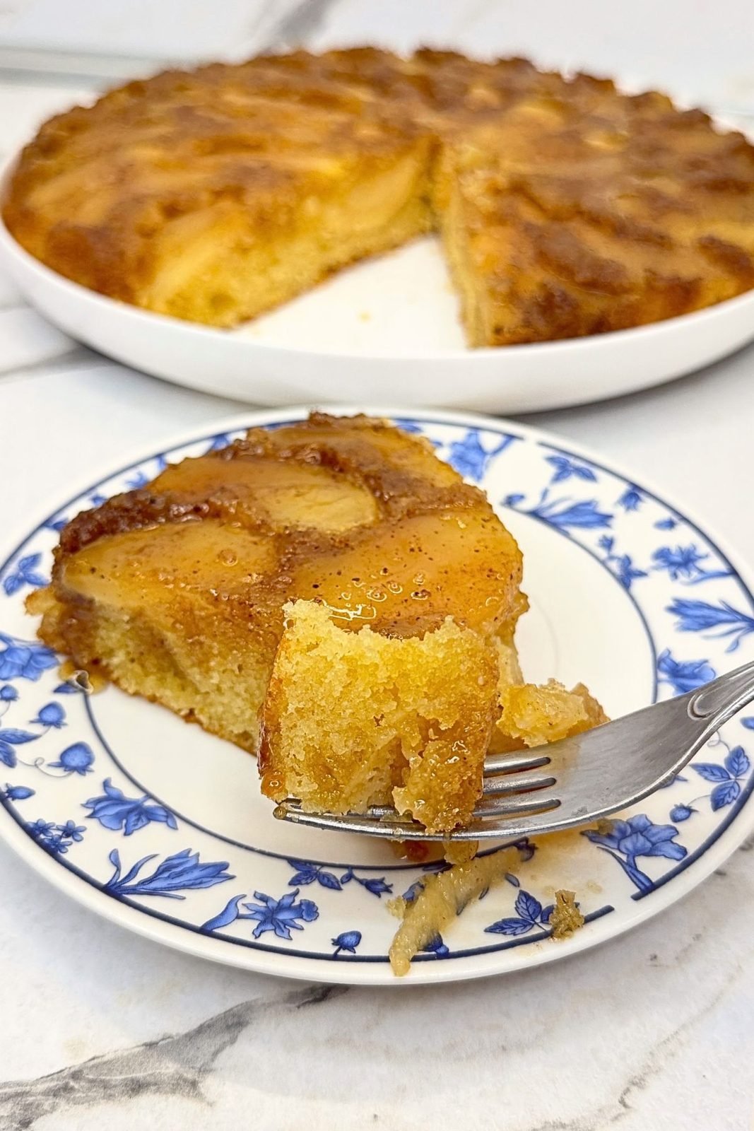 Bite of apple upside down cake on a fork, with a caramelized apple-topped slice on a blue-and-white plate.