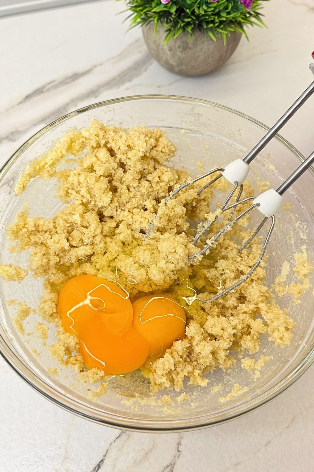 Eggs added to creamed butter and sugars in a glass mixing bowl with hand mixer beaters.
