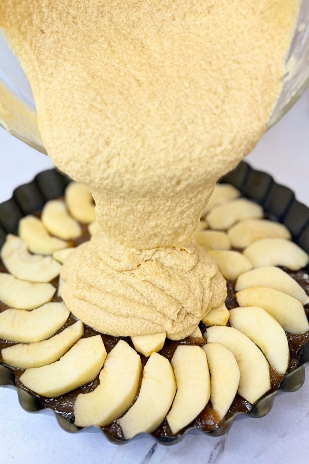 Cake batter being poured over sliced apples and caramel in a round baking pan.