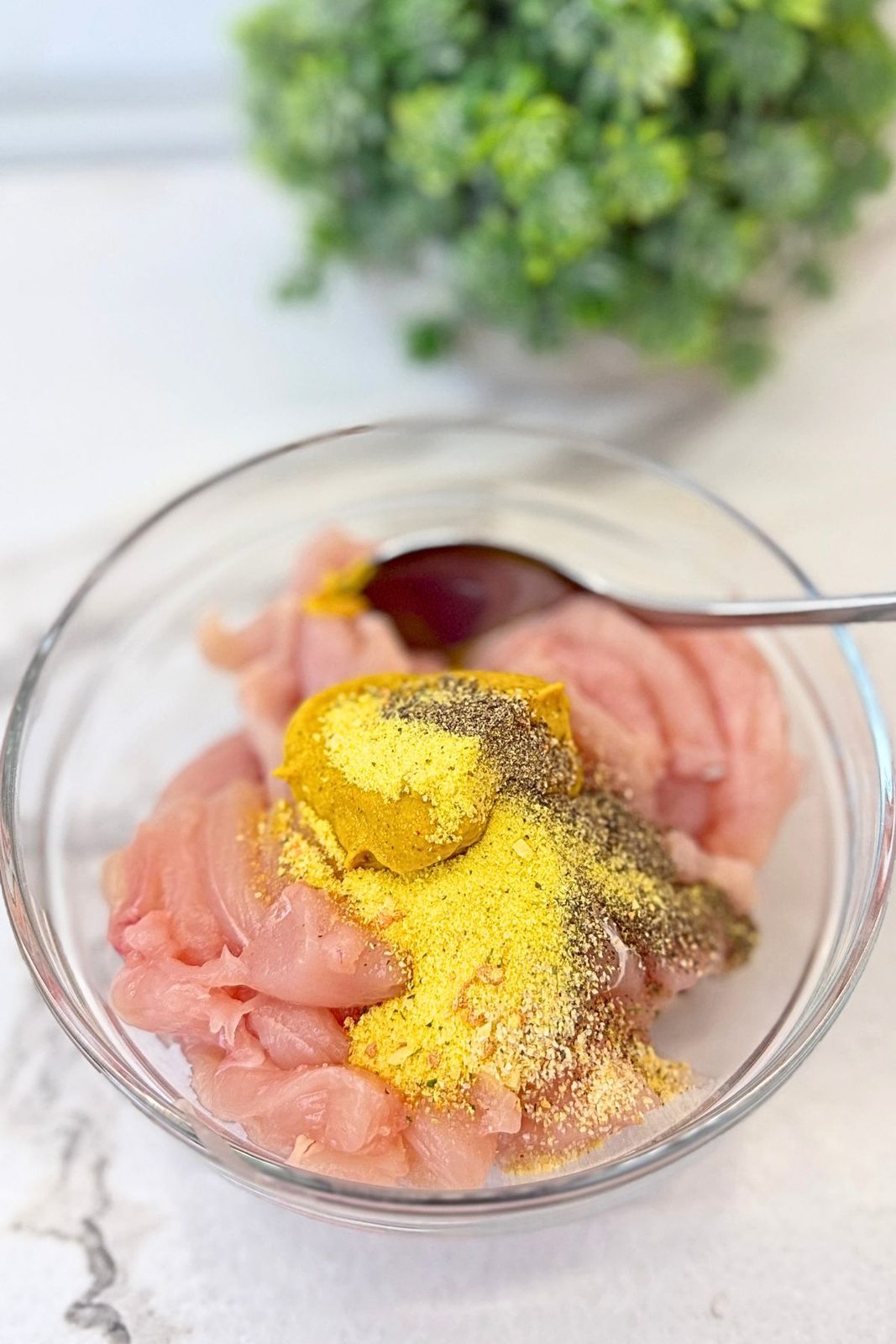 Raw chicken strips topped with mustard, black pepper, garlic powder, and seasoning mix in a glass bowl.
