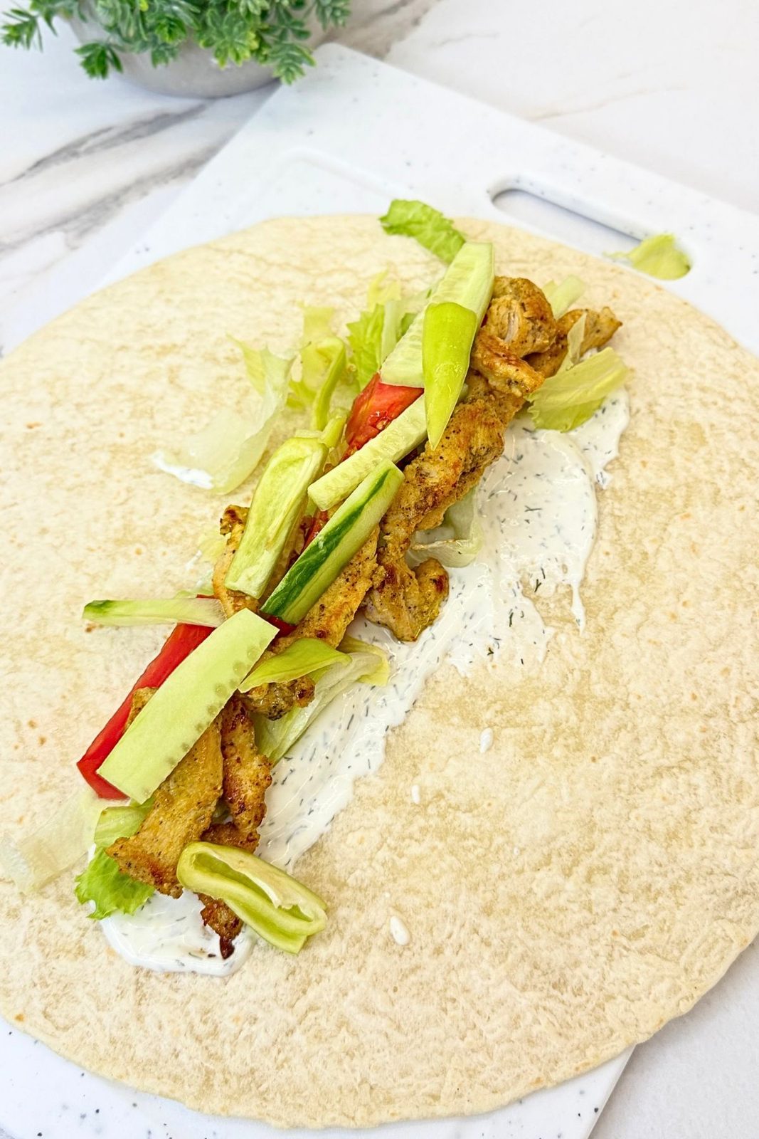 Flour tortilla spread with creamy dill sauce and topped with cooked chicken, lettuce, tomatoes, cucumber, and bell pepper before folding.
