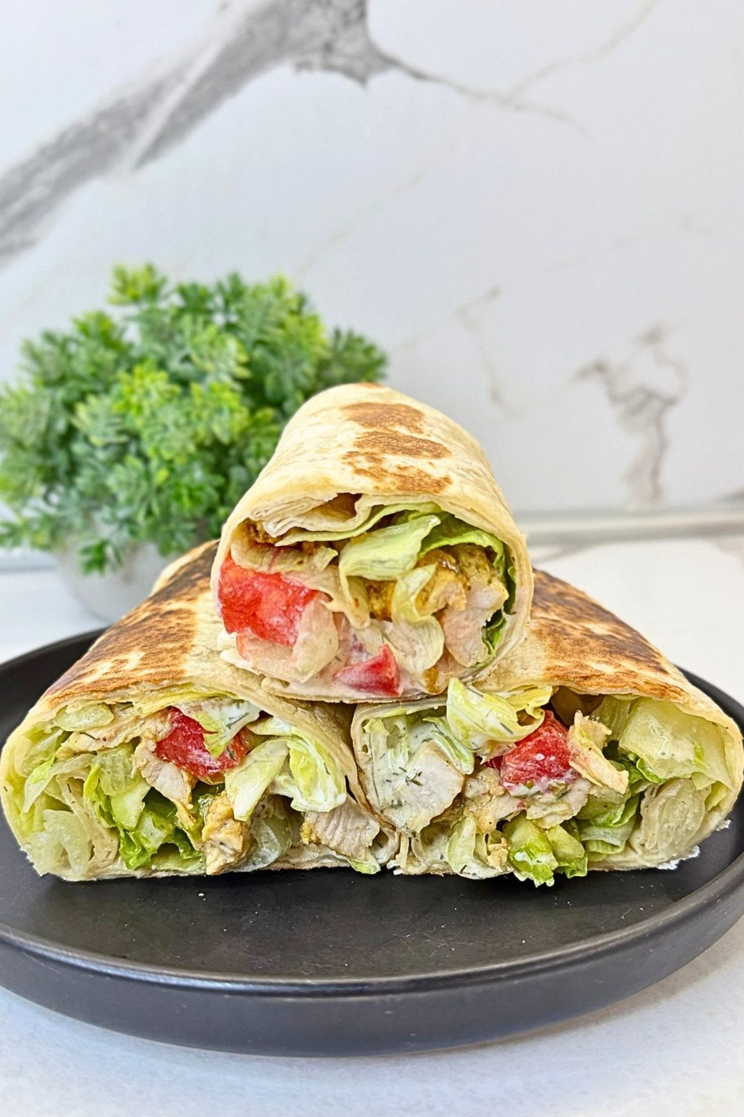 Chicken tortilla wraps with chicken, lettuce, tomatoes, cucumber, and creamy dill sauce, stacked on a black plate.

