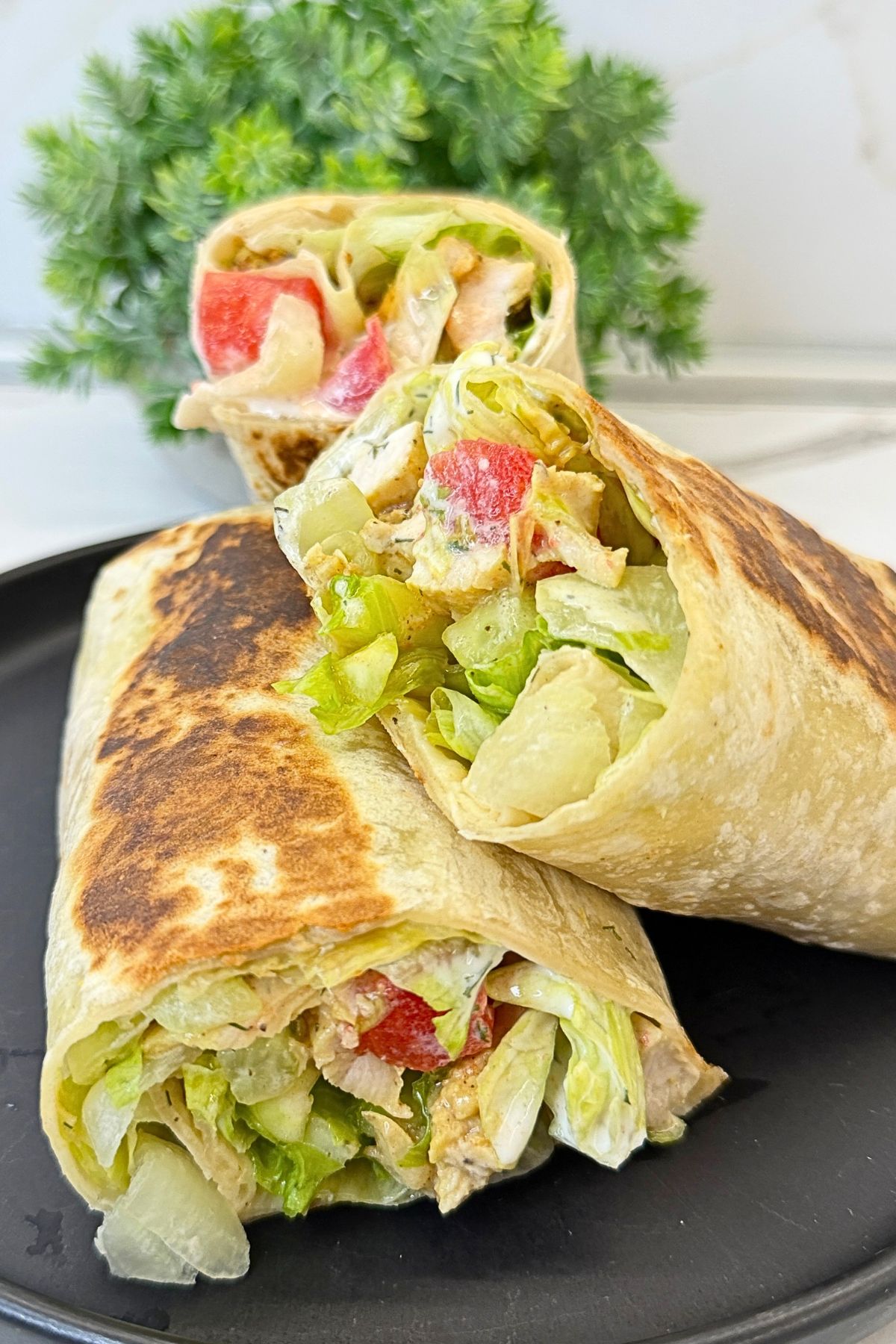 Chicken tortilla wraps stacked on a plate, filled with seasoned chicken, lettuce, tomatoes, cucumber, and creamy dill sauce.
