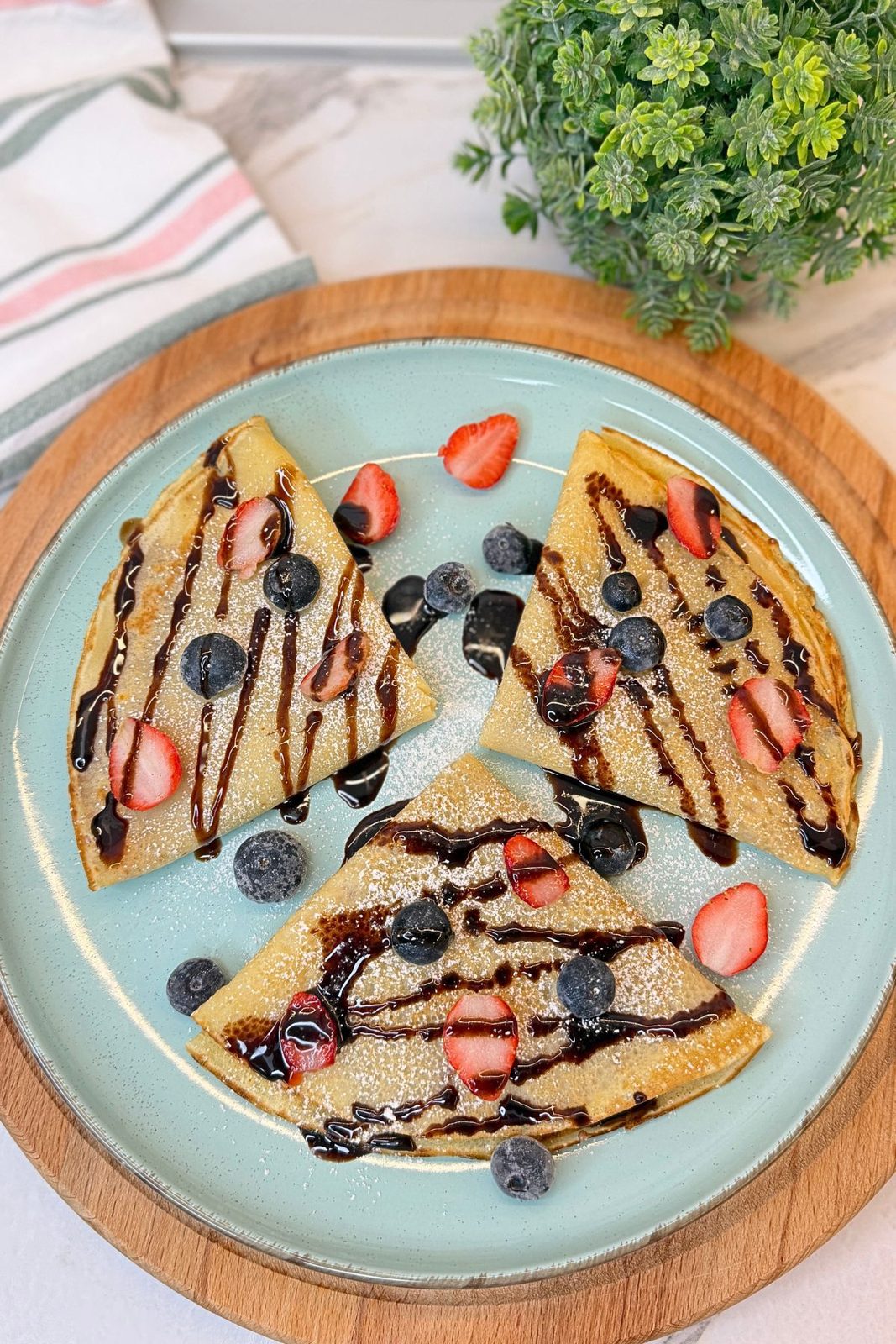Easy homemade crepes folded into triangles and topped with chocolate sauce, strawberries, blueberries, and powdered sugar