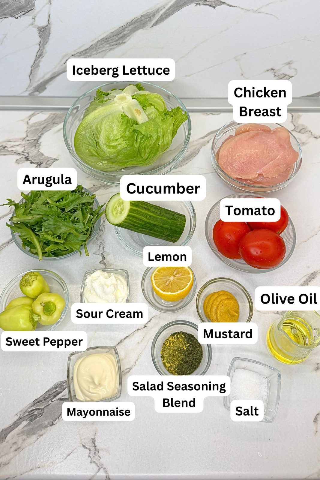 Ingredients for Easy Grilled Chicken Salad including chicken breasts, iceberg lettuce, arugula, tomatoes, cucumber, bell pepper, lemon, sour cream, mayonnaise, mustard, olive oil, and seasonings.