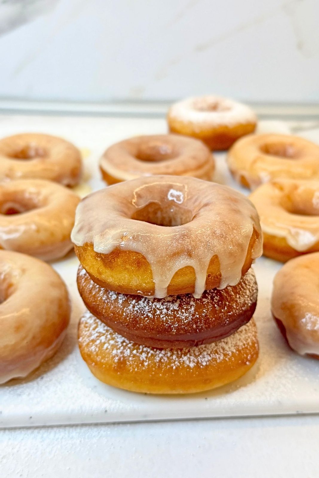 Stack of homemade donuts with vanilla glaze dripping over the top donut, surrounded by glazed donuts on a white board.