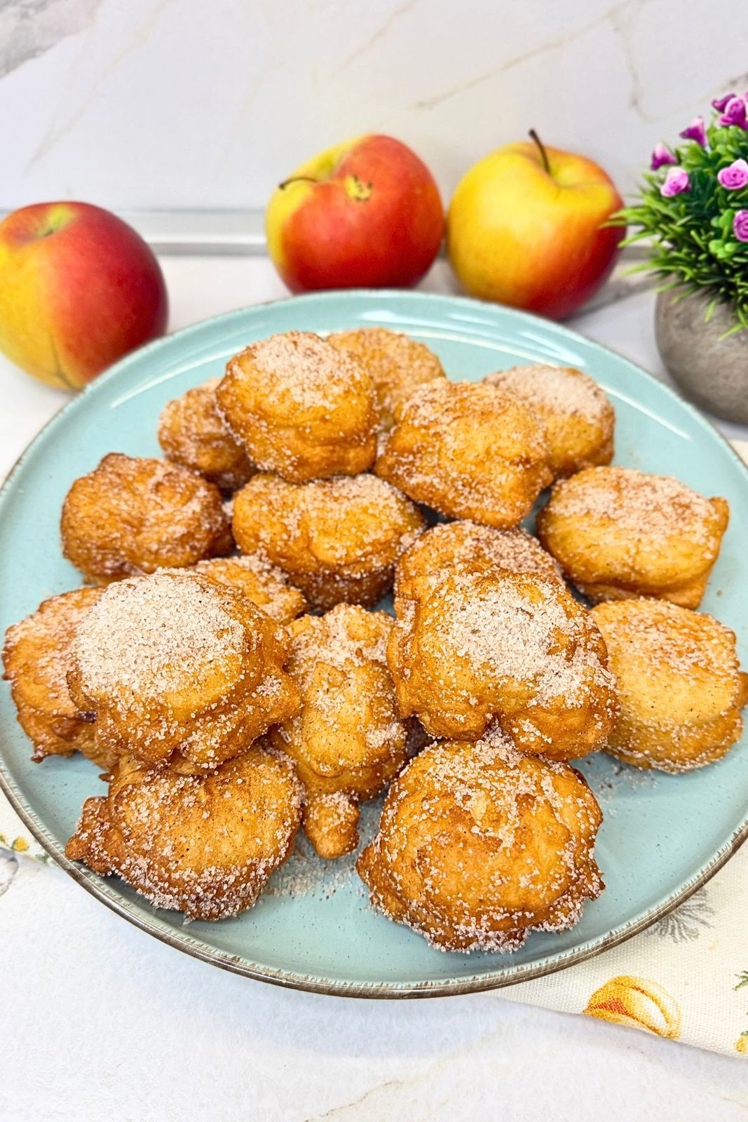 Homemade apple fritters coated in cinnamon sugar on a light blue plate with fresh apples in the background.
