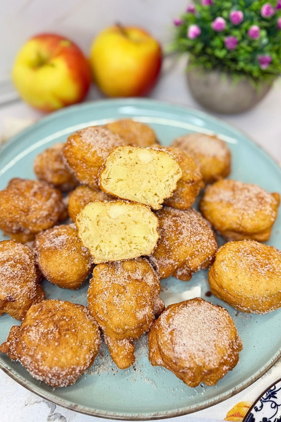 Homemade apple fritters coated in cinnamon sugar on a blue plate, with a halved fritter showing the soft center and apple pieces.