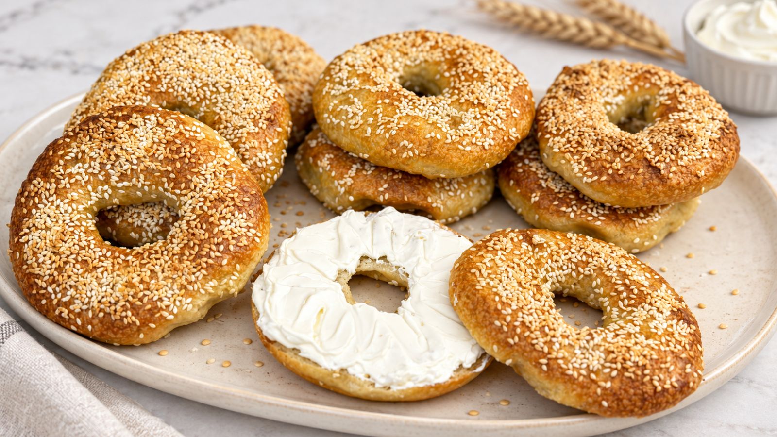 Homemade sesame bagels stacked on a ceramic platter, with a sliced bagel spread with cream cheese in the foreground.