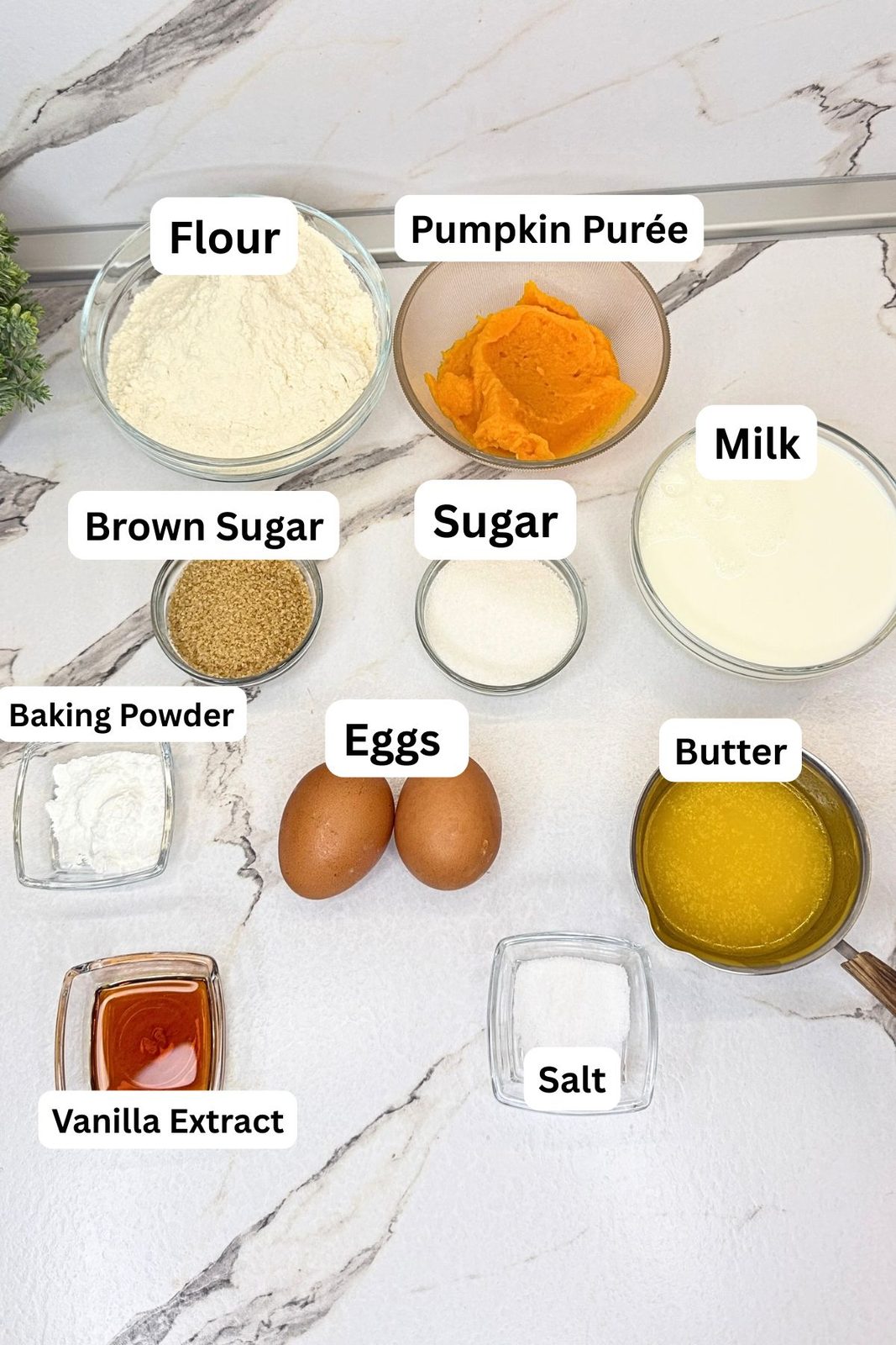 Ingredients for pumpkin pancakes in labeled bowls, including flour, pumpkin purée, milk, brown sugar, granulated sugar, baking powder, eggs, melted butter, vanilla extract, and salt.