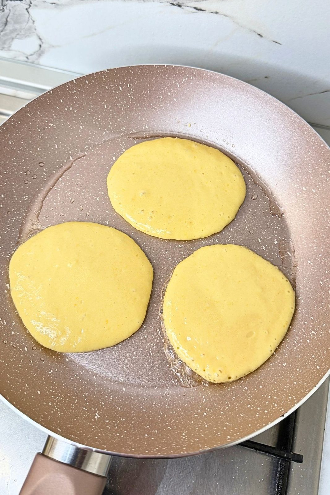 Three pumpkin pancakes cooking in a nonstick skillet with small bubbles forming on the surface.