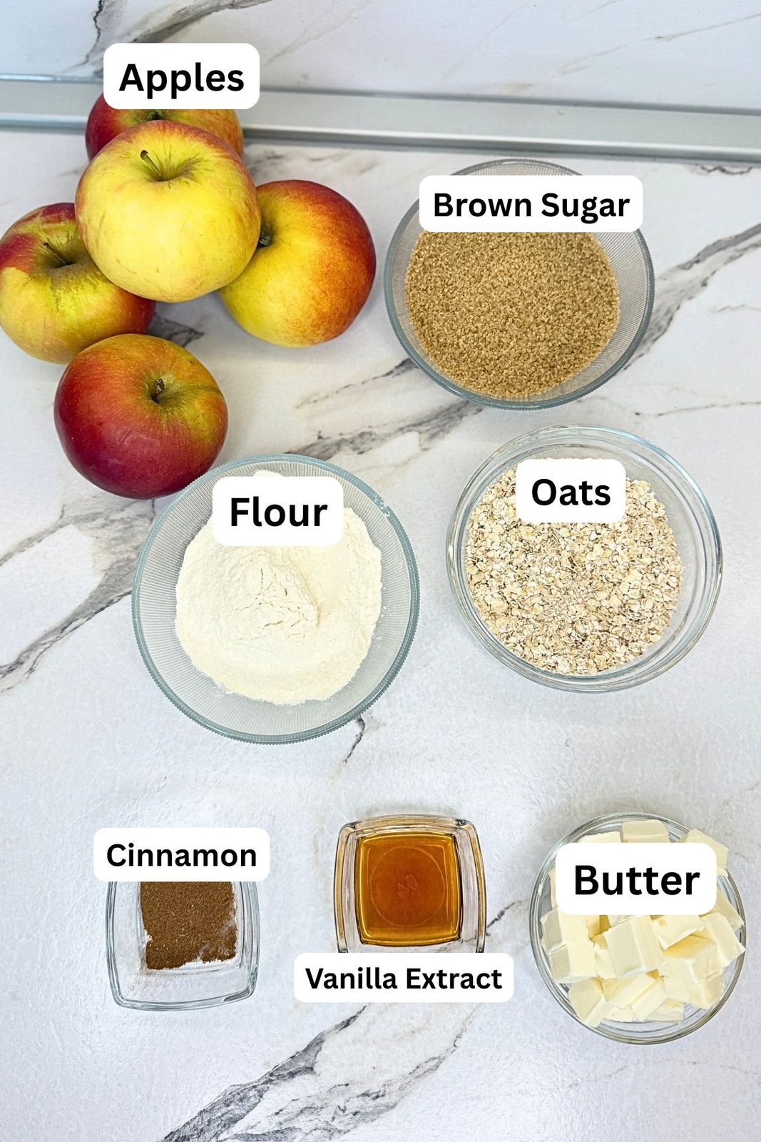 Apple crisp ingredients on a marble countertop, including apples, brown sugar, oats, flour, cinnamon, vanilla extract, and cubed butter in labeled bowls.