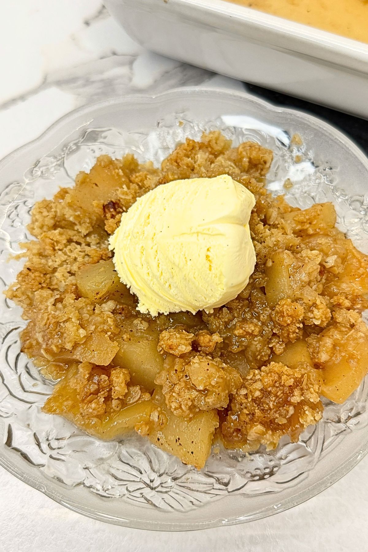 Apple crisp with buttery oat topping and tender cinnamon apples, served with vanilla ice cream, with a baking dish in the background.