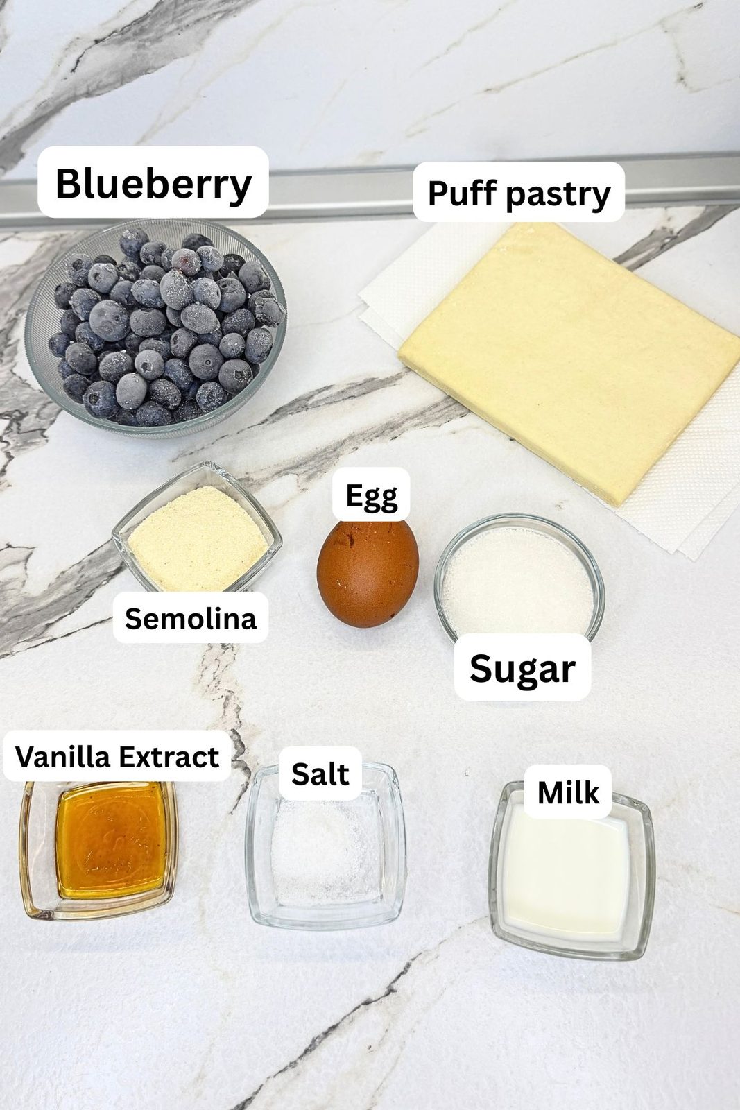 Ingredients for blueberry turnovers: blueberries, puff pastry, semolina, sugar, egg, vanilla extract, salt, and milk arranged on a marble countertop.