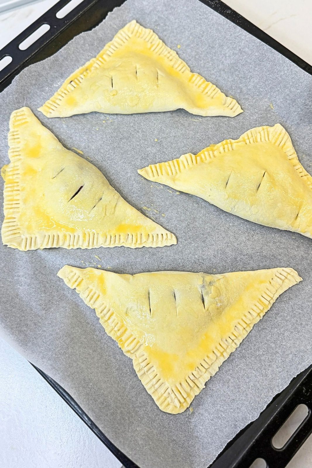Unbaked blueberry turnovers with fork-crimped edges, egg-washed tops, and steam vents on a parchment-lined baking sheet.