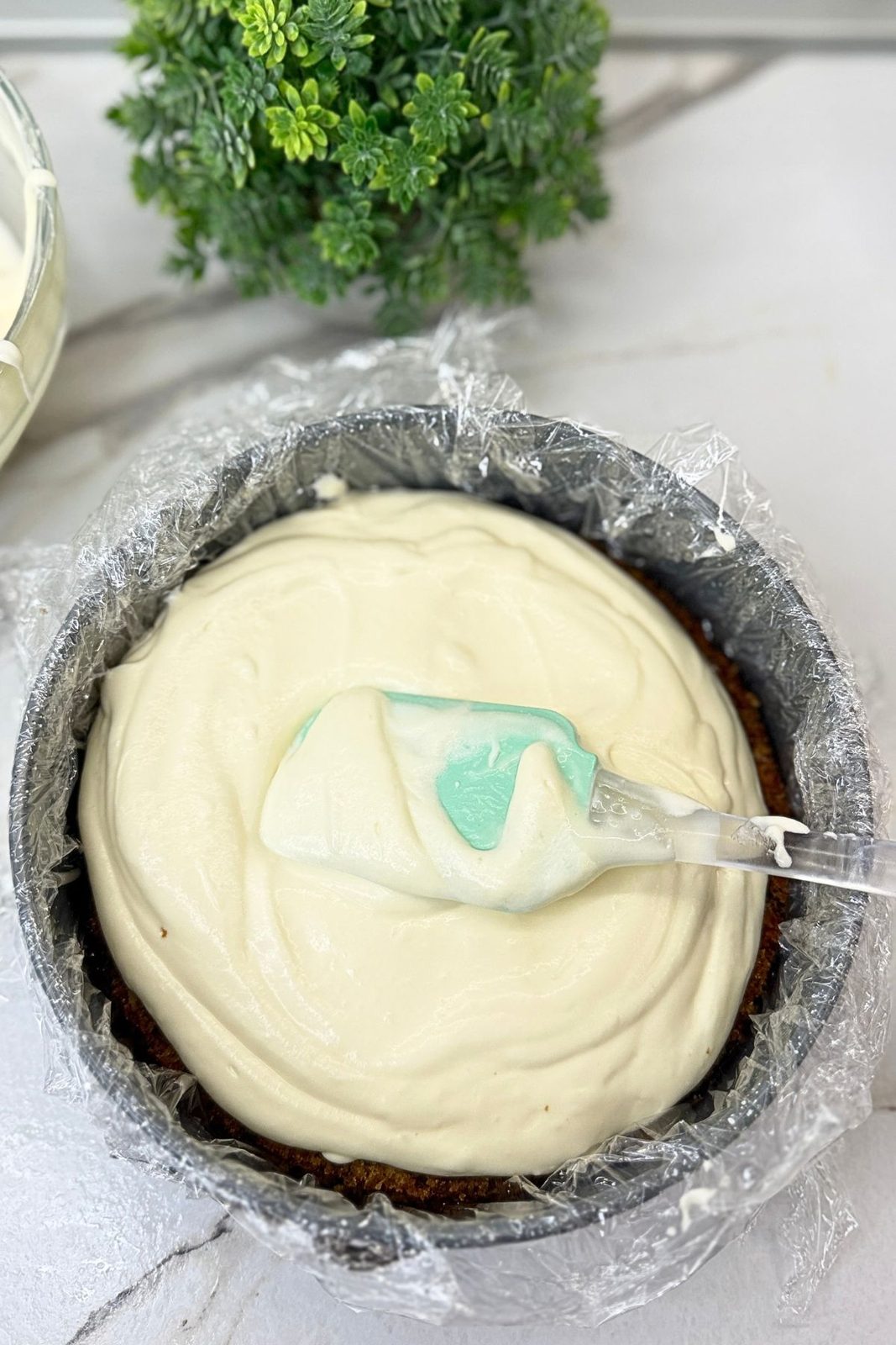 Cream cheese frosting spread over the first carrot cake layer in a pan lined with plastic wrap.