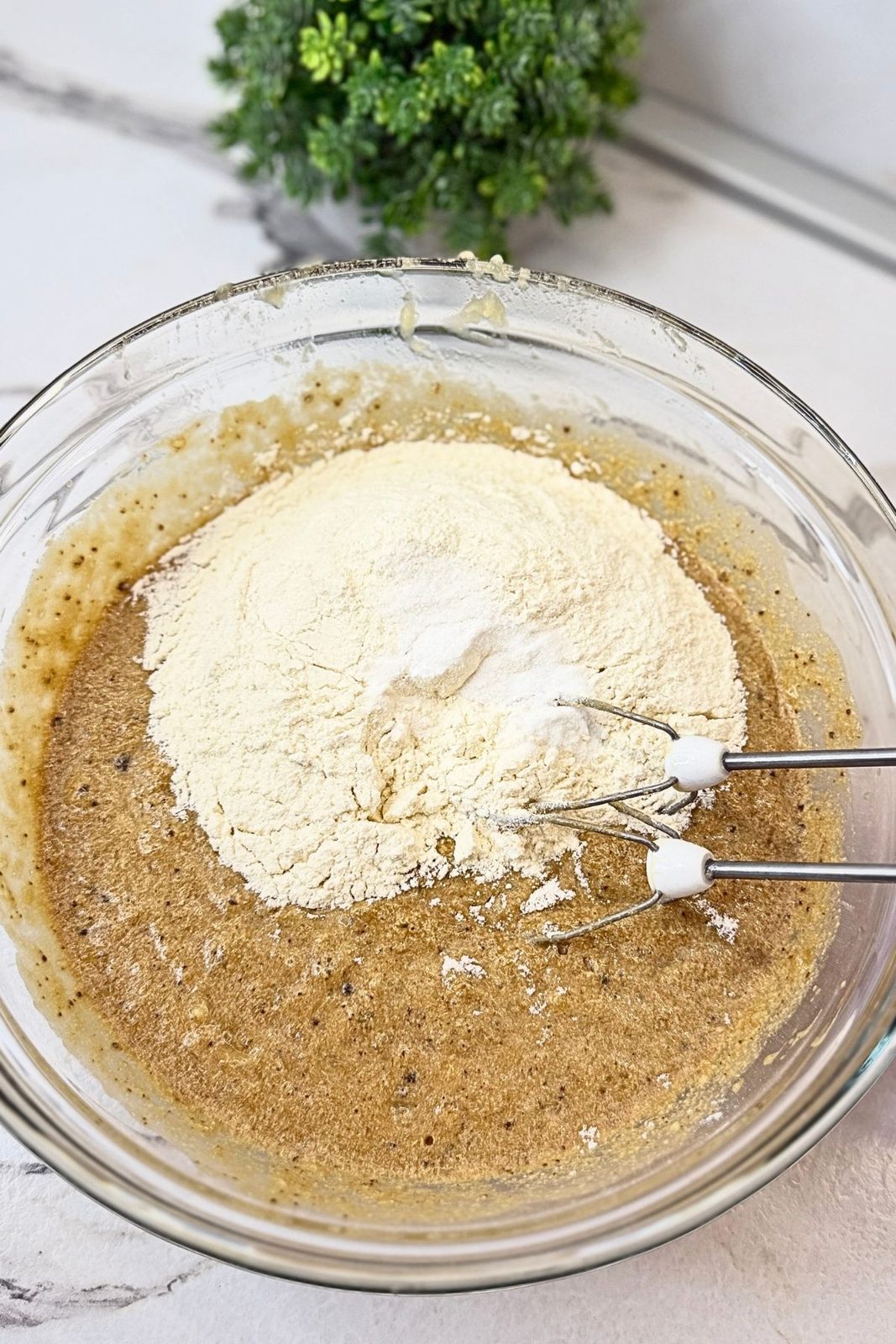 Flour mixture added to the wet banana bread ingredients in a glass mixing bowl.
