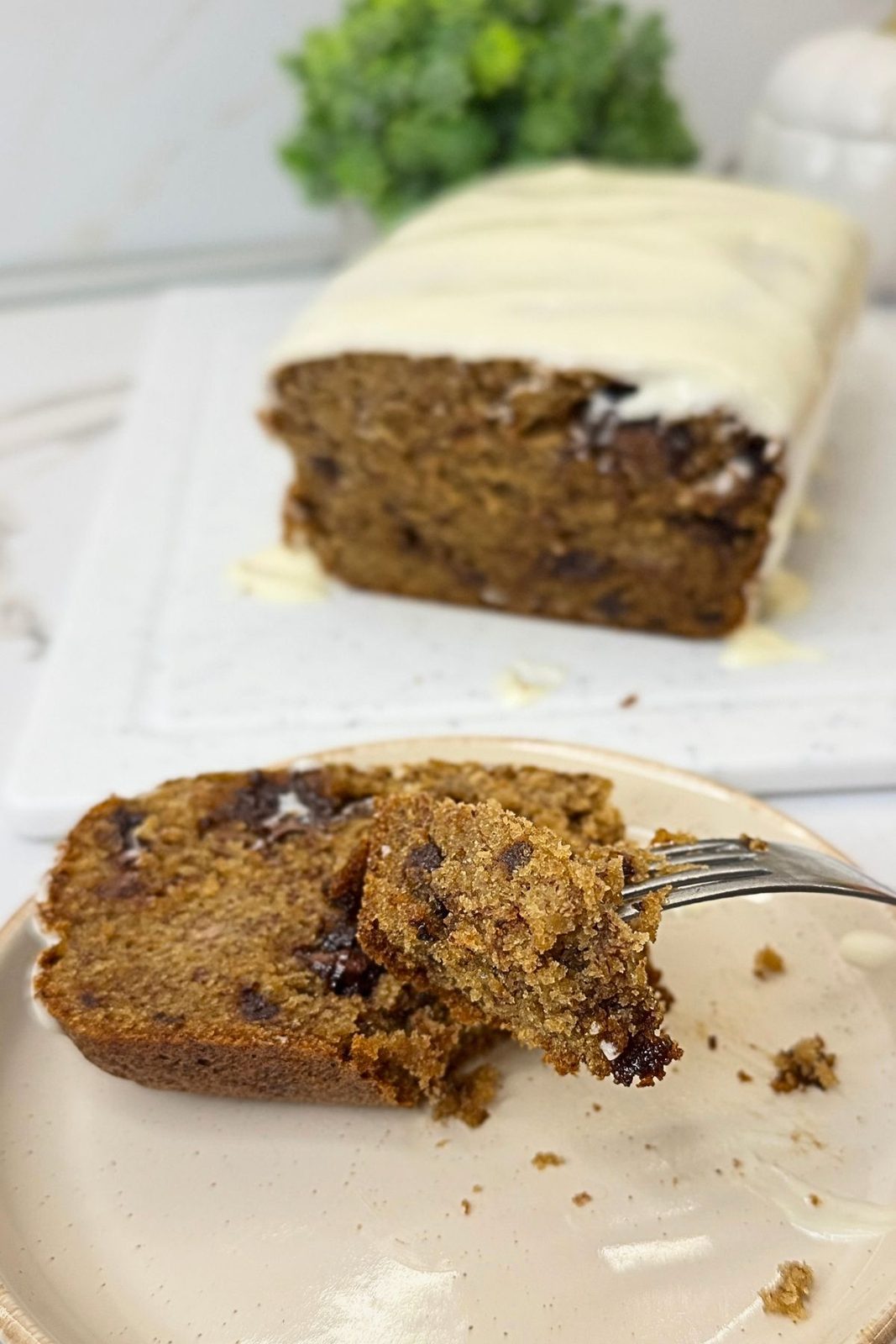 A bite of chocolate chip banana bread on a fork, with a slice on a plate and the cream cheese frosted loaf in the background.