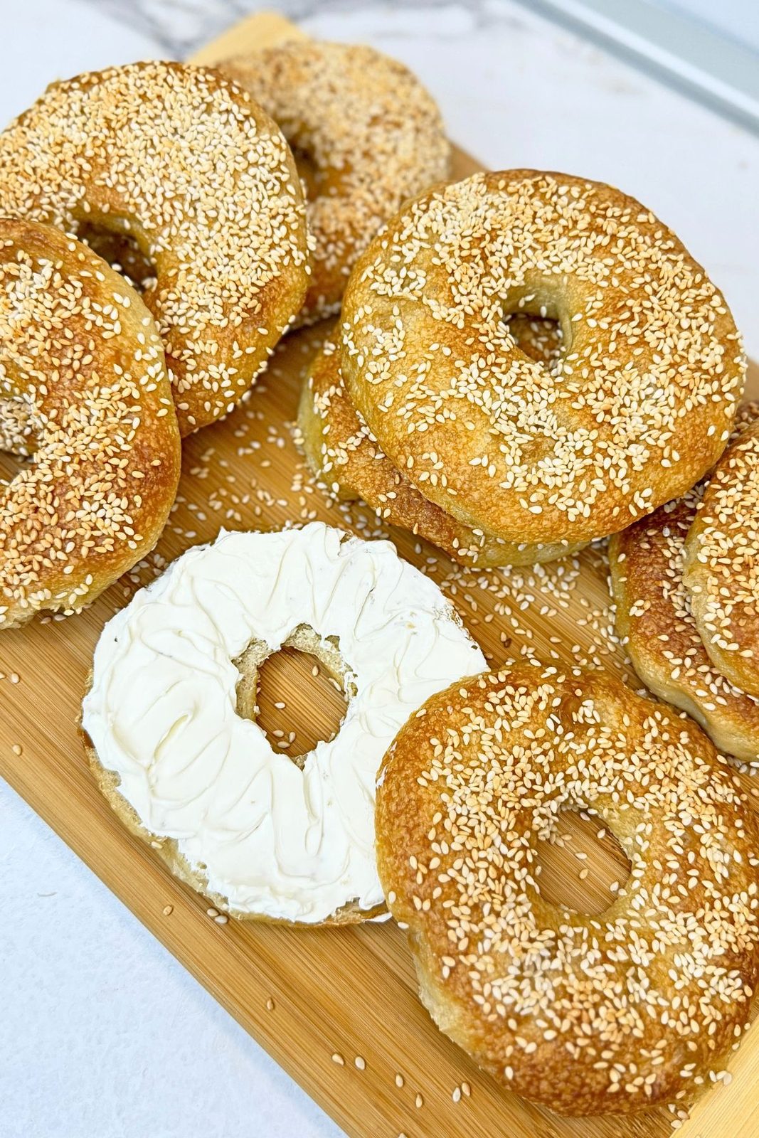 Homemade bagels topped with sesame seeds on a wooden board, with a sliced half spread with cream cheese in the foreground.