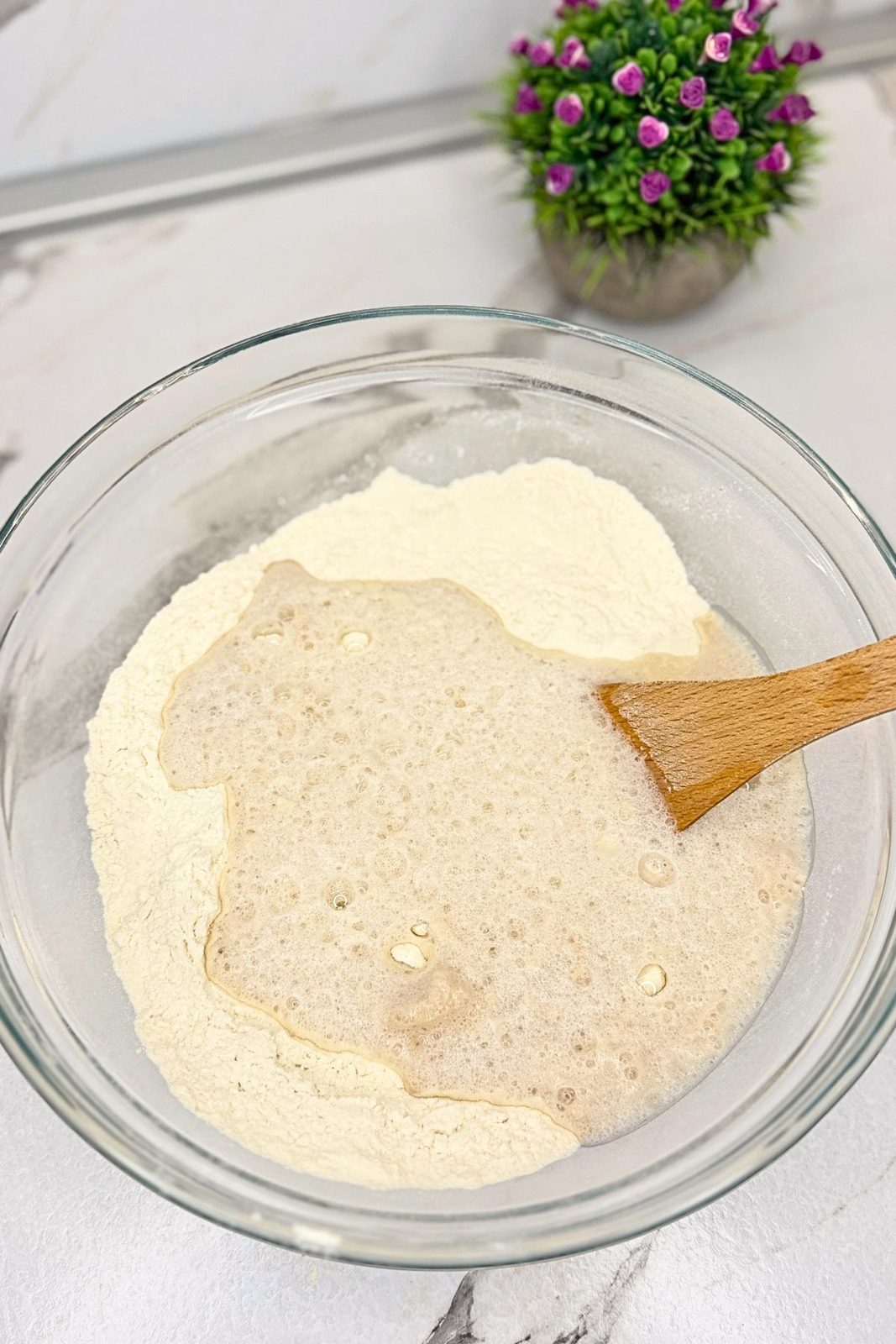 Flour and activated yeast mixture in a glass bowl with a wooden spoon, ready to mix into bagel dough.