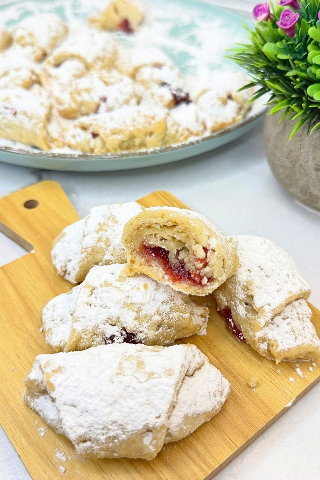 Jam filled crescent cookies dusted with powdered sugar on a wooden board, with a cut cookie showing cranberry filling and a platter of cookies in the background.