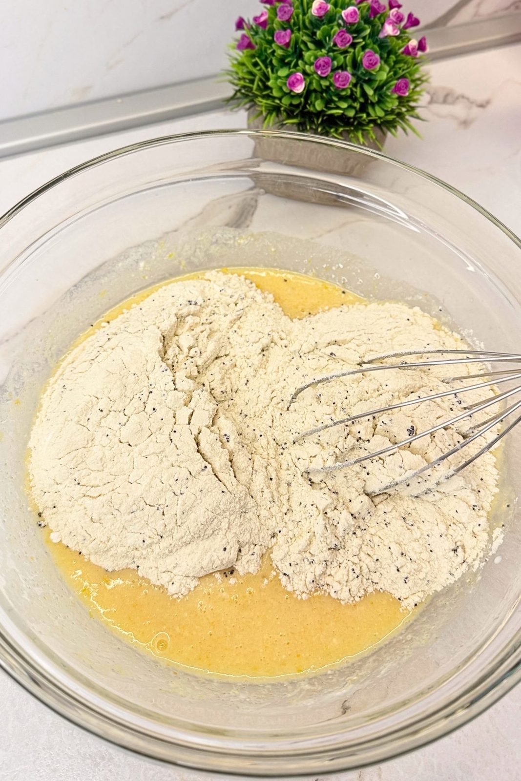 Flour mixture with poppy seeds added to the wet ingredients in a glass mixing bowl.