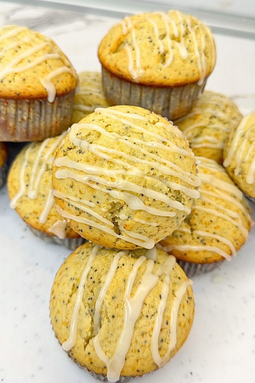 A halved lemon poppy seed muffin showing its soft interior, surrounded by whole muffins topped with lemon glaze.