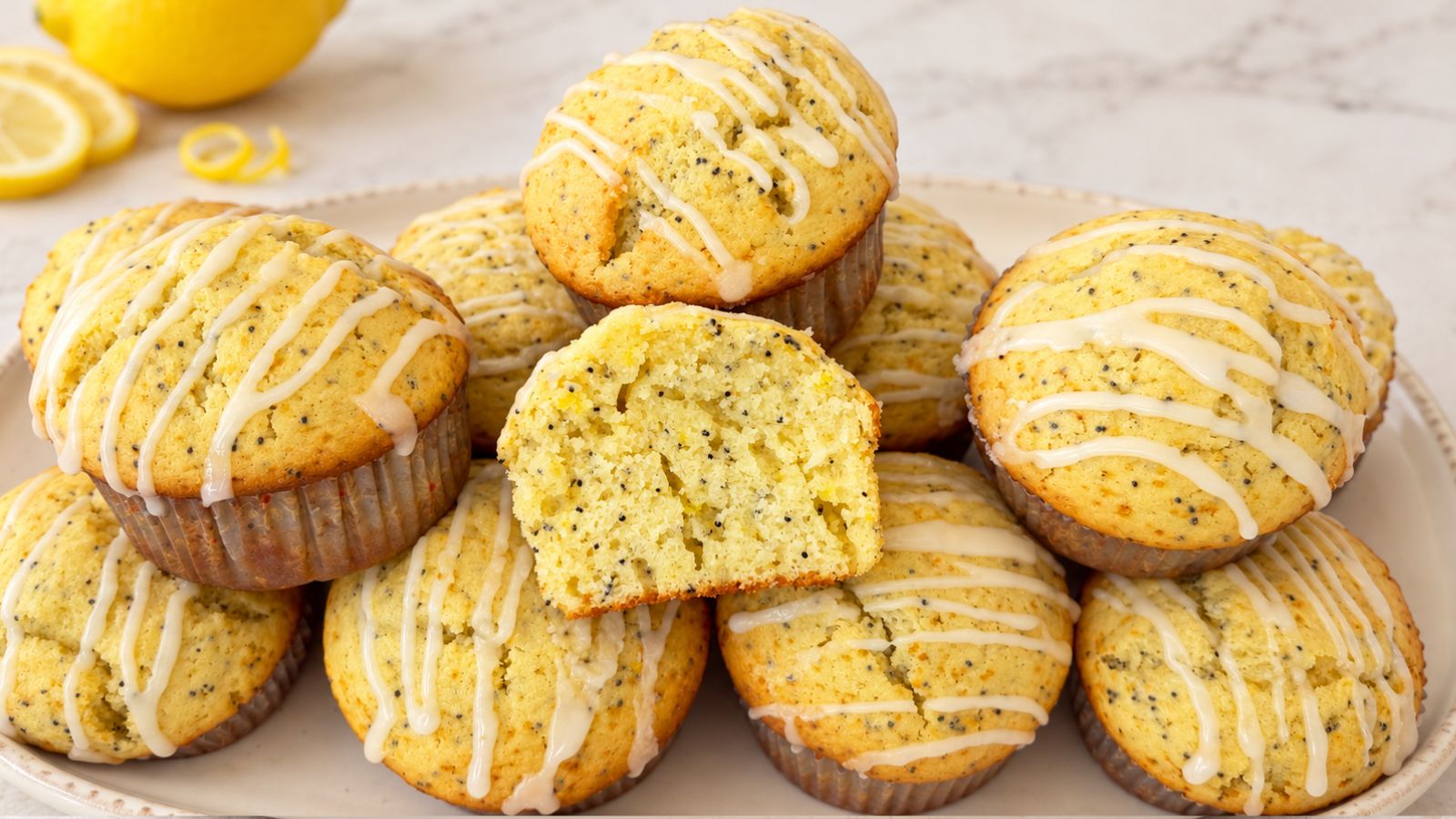 Lemon poppy seed muffins drizzled with lemon glaze on a platter, with a halved muffin showing its soft interior and fresh lemons in the background.