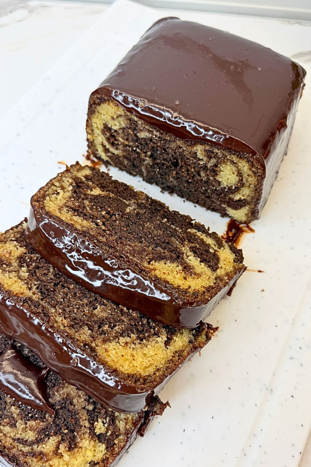 Marble loaf cake with chocolate ganache and sliced pieces showing vanilla and chocolate swirls on a cream-colored platter.