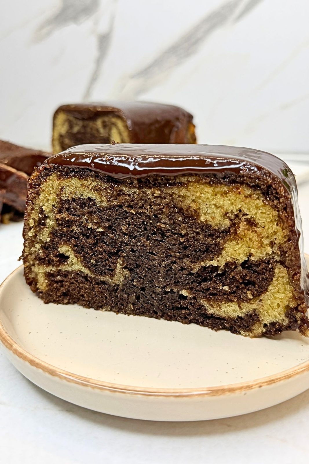 Close-up of marble loaf cake with vanilla and chocolate swirls, topped with glossy chocolate ganache on a beige plate.