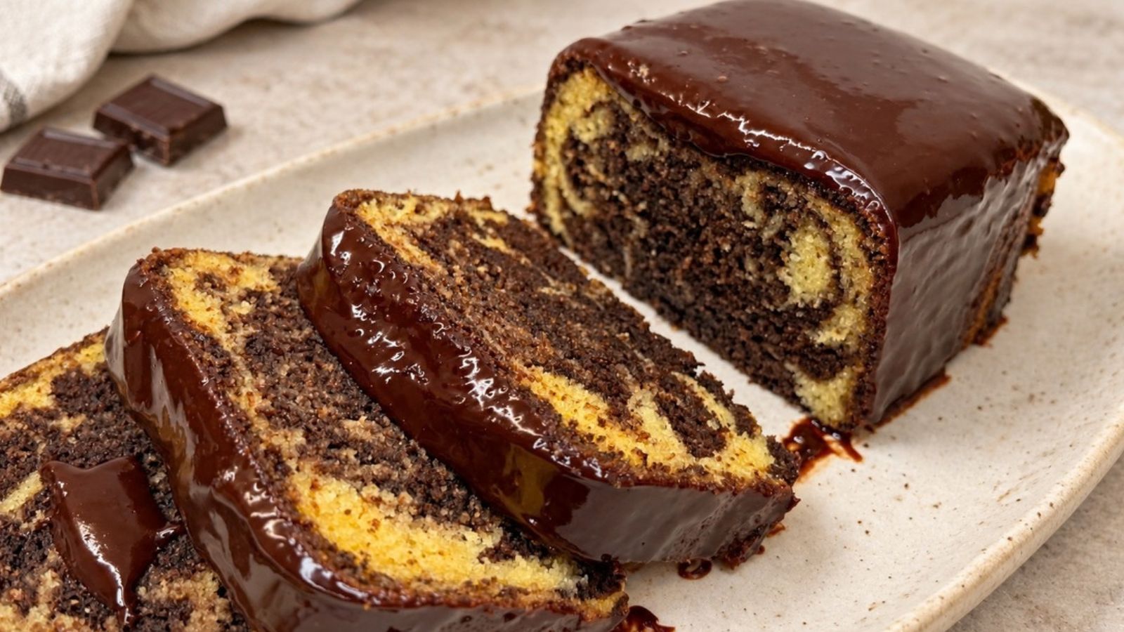 Marble loaf cake with chocolate ganache and sliced pieces showing vanilla and chocolate swirls on a cream-colored platter.