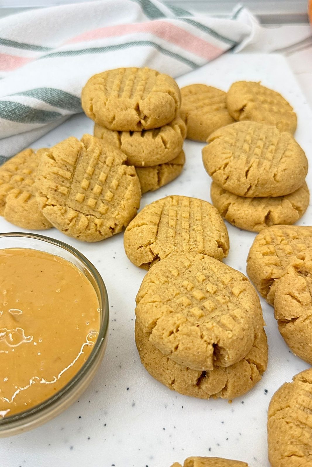 Peanut butter cookies with crisscross fork marks stacked on a white surface beside a bowl of creamy peanut butter.