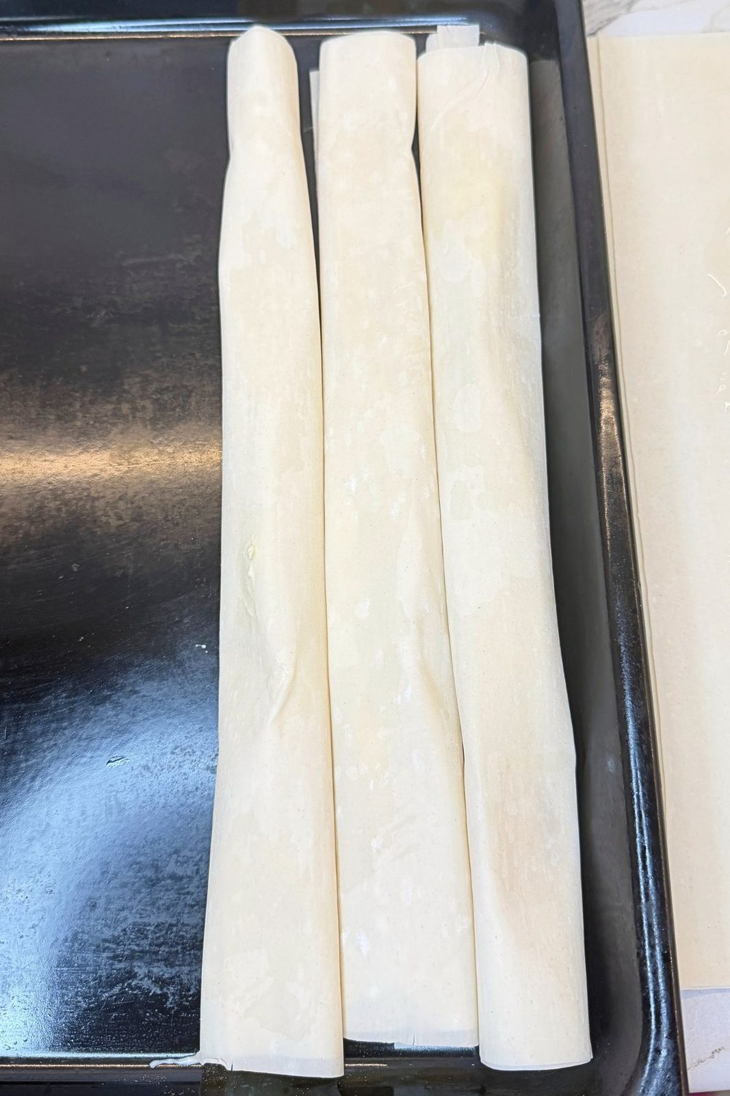 Three unbaked phyllo cheese rolls arranged side by side in a black baking pan.