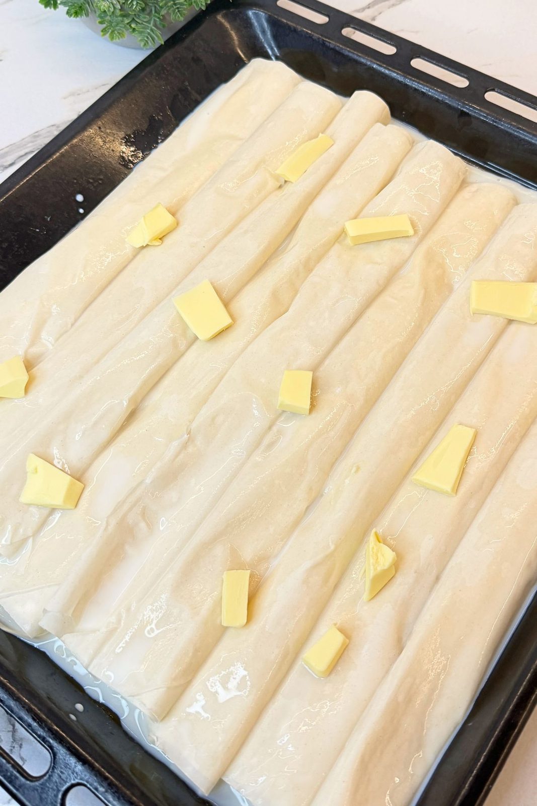Unbaked phyllo cheese rolls topped with milk and small pieces of butter, ready for the oven.