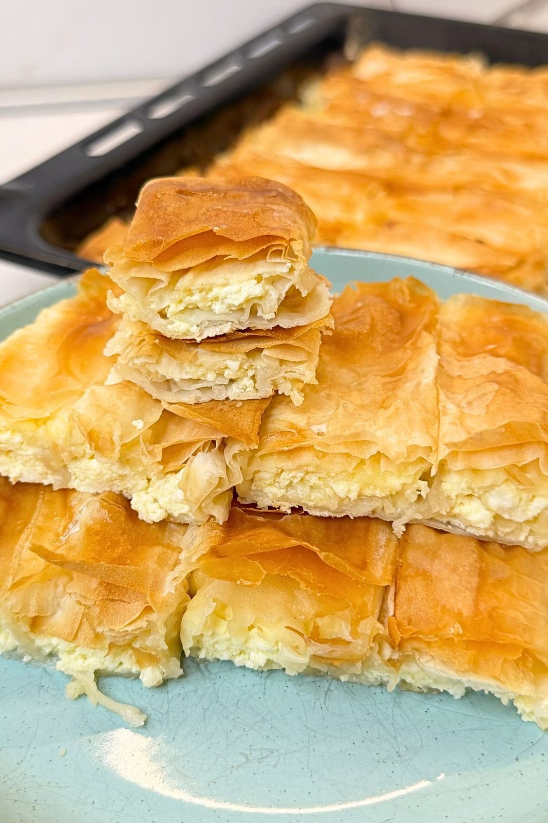 Flaky phyllo cheese rolls stacked on a turquoise plate, showing the creamy cheese filling, with a baking tray in the background.