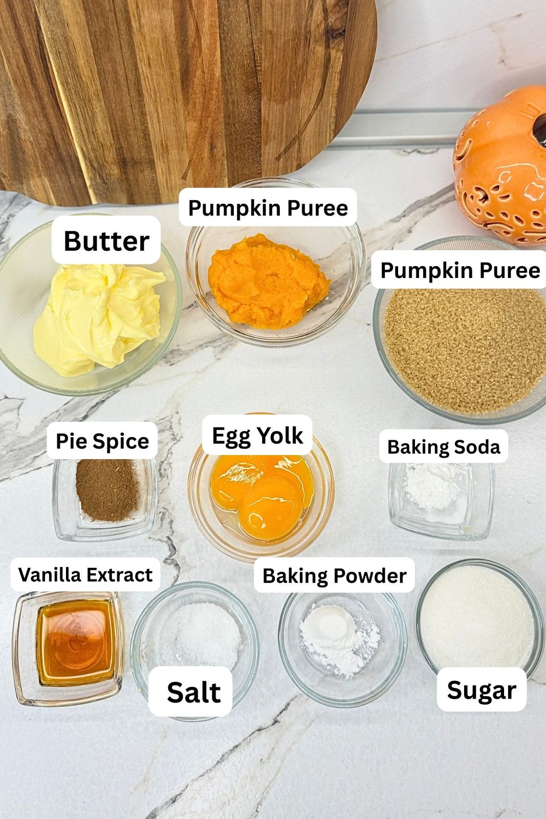 Ingredients for pumpkin cookies in labeled bowls, including butter, pumpkin purée, brown sugar, egg yolks, pumpkin pie spice, vanilla, baking soda, baking powder, salt, and sugar.

