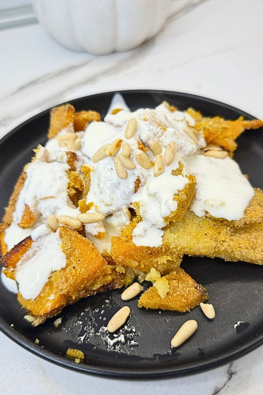 Roasted pumpkin and potatoes with golden cornmeal coating, topped with garlic yogurt sauce and pine nuts on a black plate.