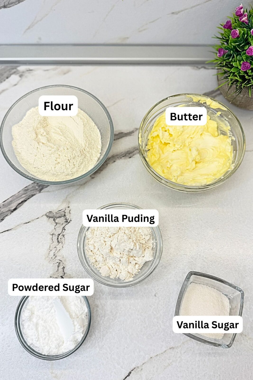 Ingredients for vanilla crescent cookies: all-purpose flour, butter, vanilla pudding mix, powdered sugar, and vanilla sugar in glass bowls on a marble countertop.