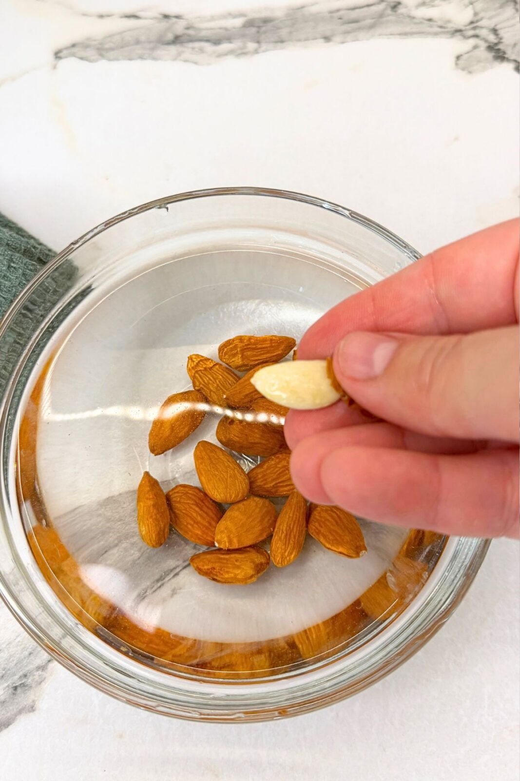 Skin-on almonds cooling in a glass bowl of cold water beside a small bowl with two peeled almonds.