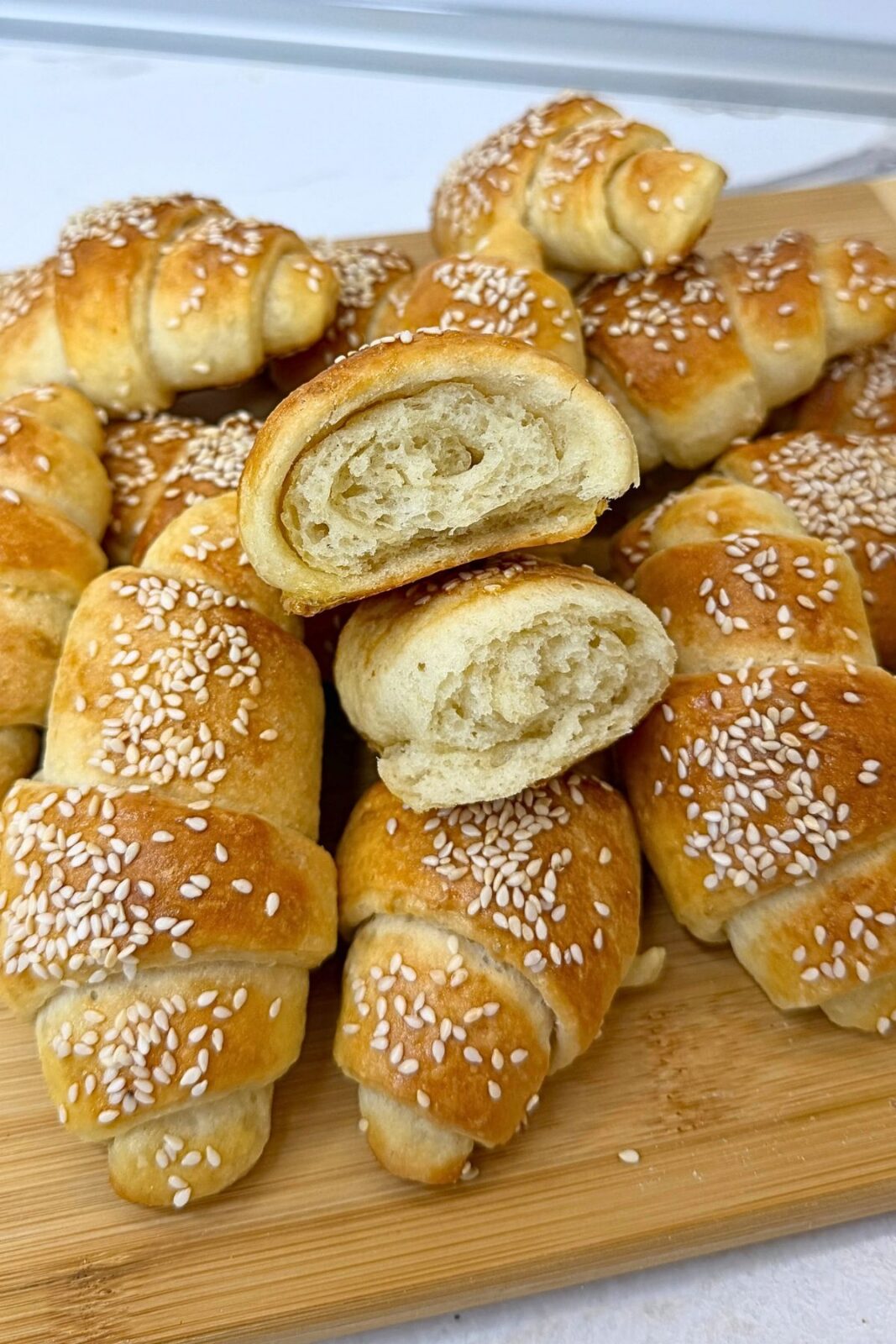 Homemade crescent rolls topped with sesame seeds on a wooden board, with two cut halves stacked in the center showing the soft interior.
