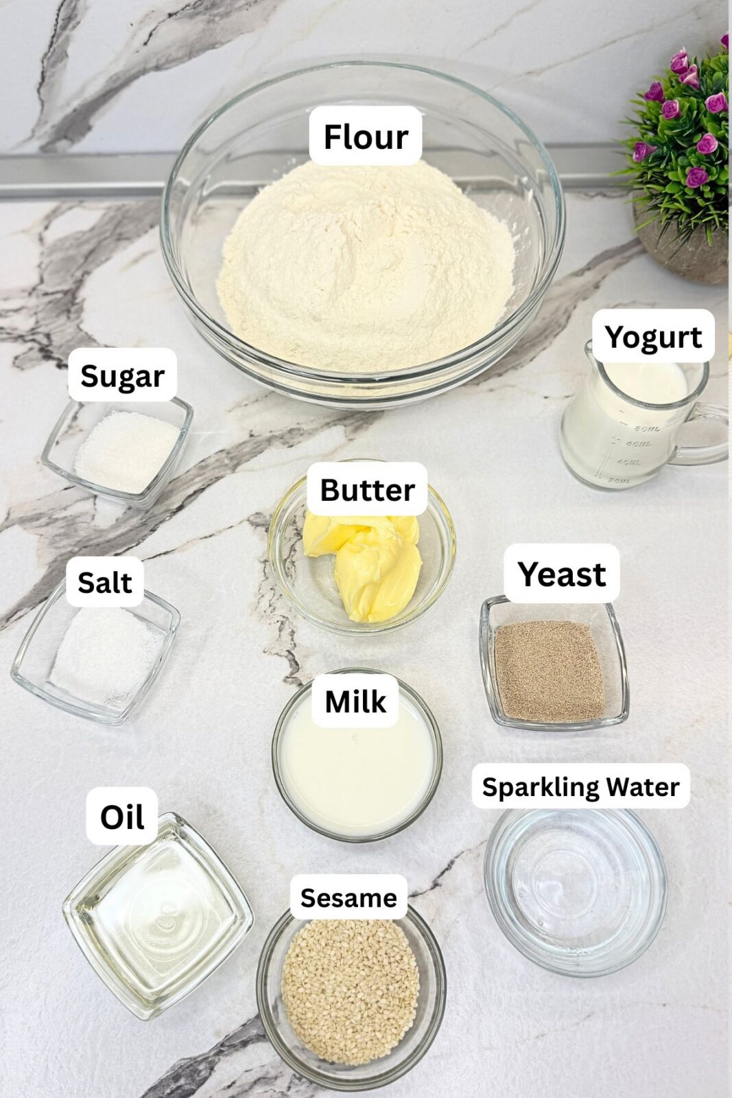 Ingredients for homemade crescent rolls arranged on a marble countertop: flour, sugar, salt, yeast, butter, oil, milk, plain yogurt, sparkling water, and sesame seeds.
