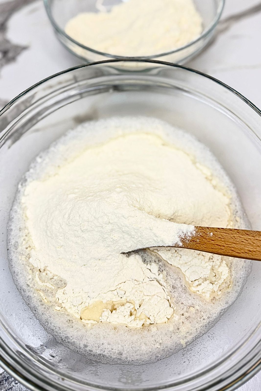 Flour added to the yeast mixture in a glass bowl with a wooden spoon.
