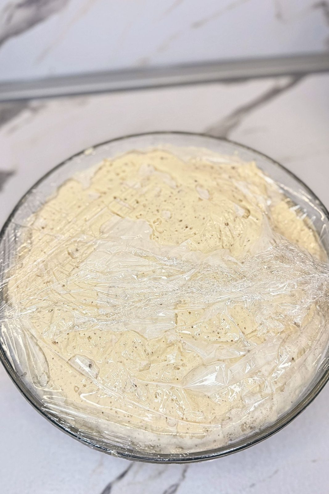 Risen sandwich bread dough in a glass bowl covered with plastic wrap.
