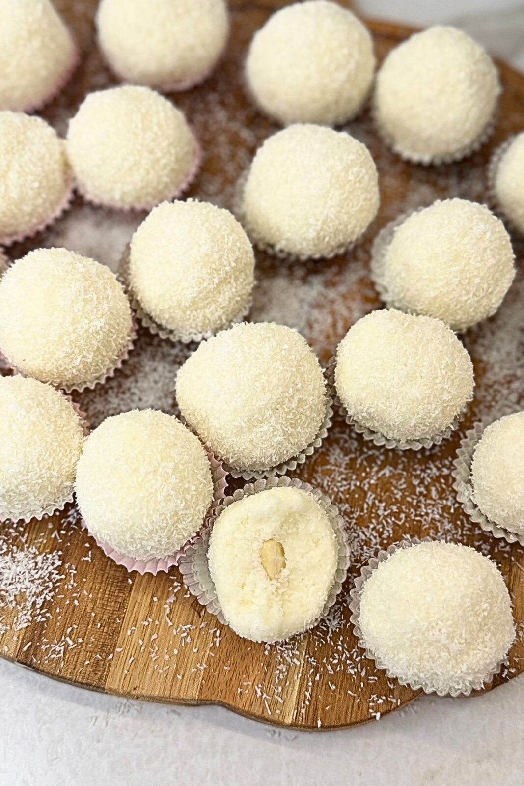 Raffaello coconut balls in paper cups on a wooden board, with one cut open to show the creamy filling and whole almond center.