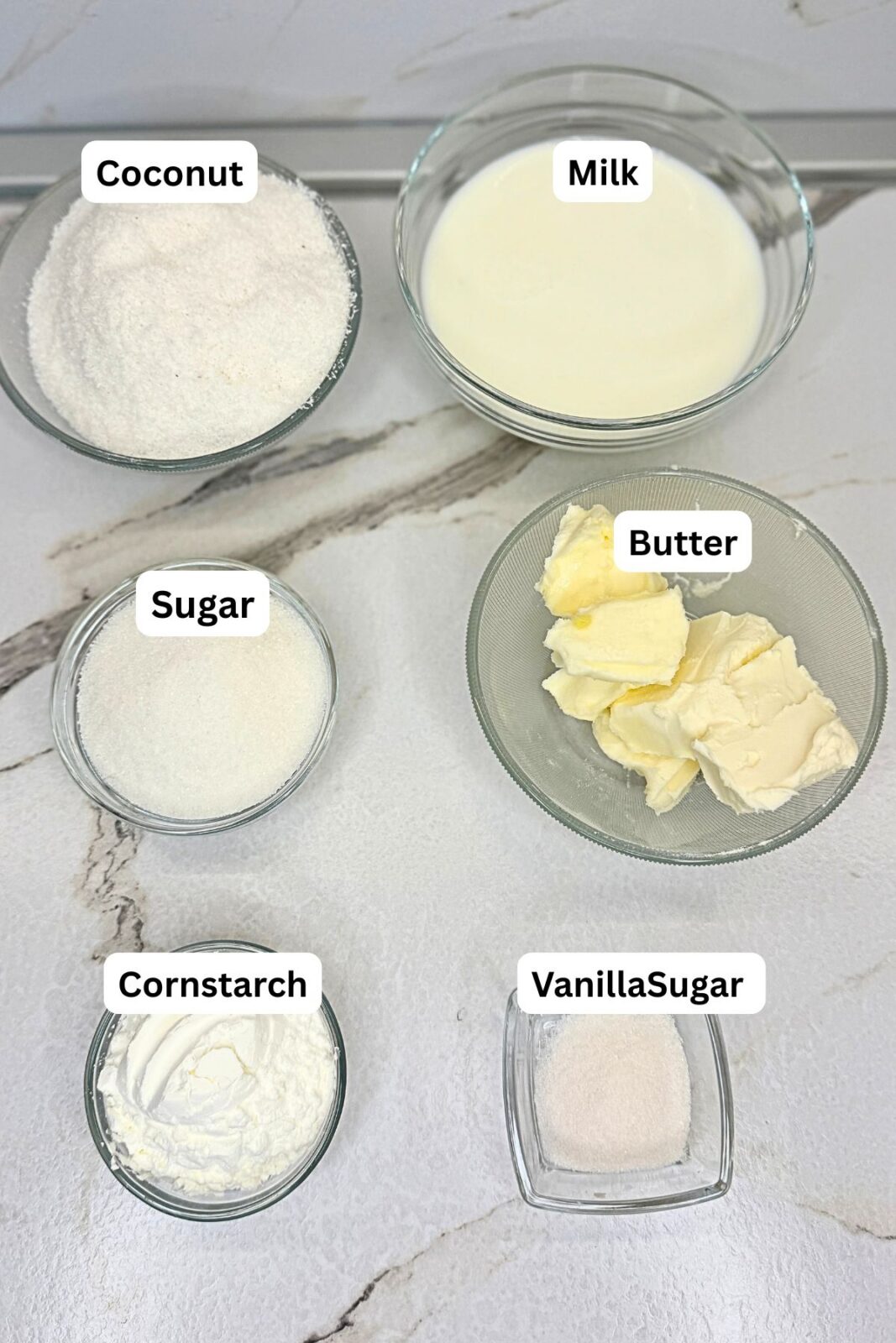 Ingredients for Raffaello coconut balls: milk, sugar, butter, desiccated coconut, cornstarch, and vanilla sugar in bowls on a marble countertop.